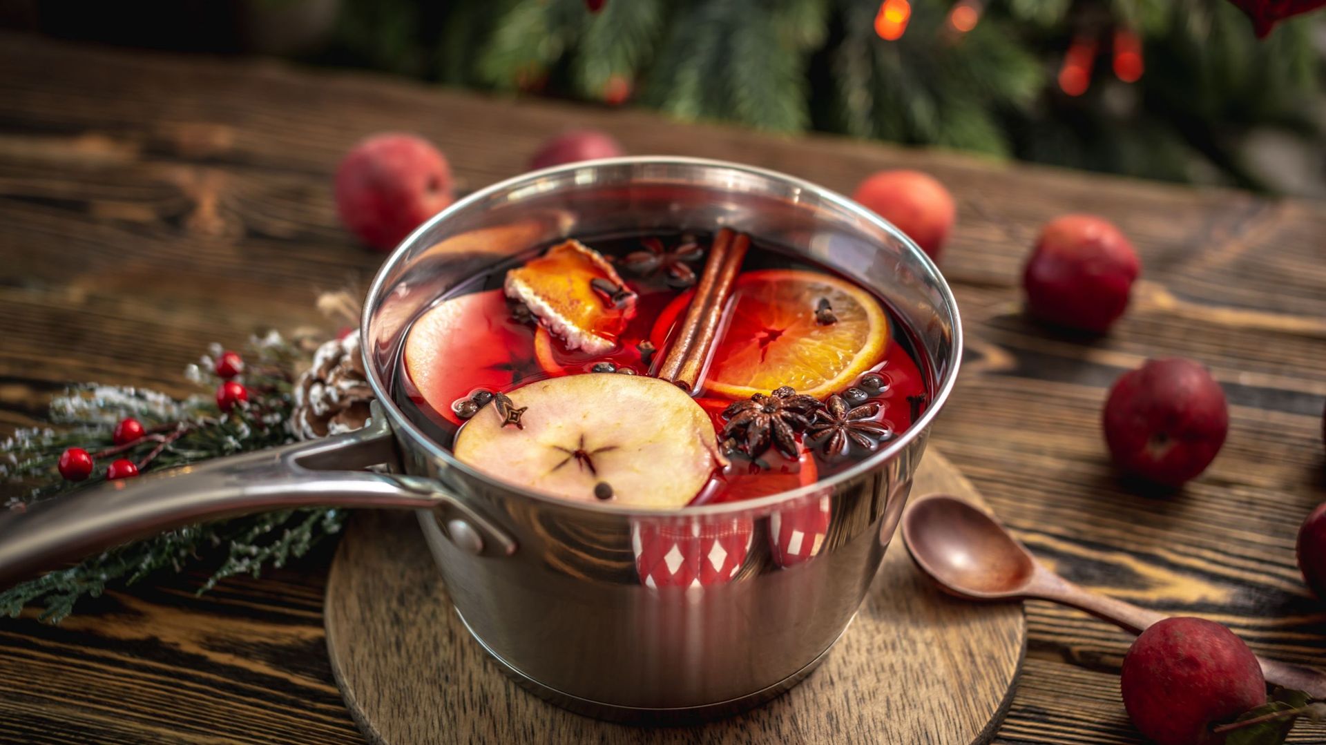 Pot with spicy hot mulled wine on a wooden table with fruit and a Christmas tree in the background. Concept of a cozy festive atmosphere, New Year mood