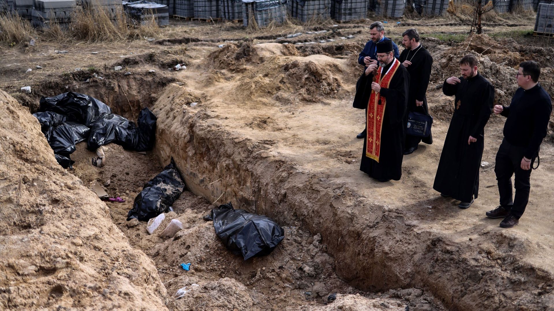 Des prêtres prient devant des sacs mortuaires dans une fosse commune située sur le terrain entourant l’église Saint-André à Boutcha, le 7 avril 2022, dans le cadre de l’invasion militaire lancée par la Russie en Ukraine.
