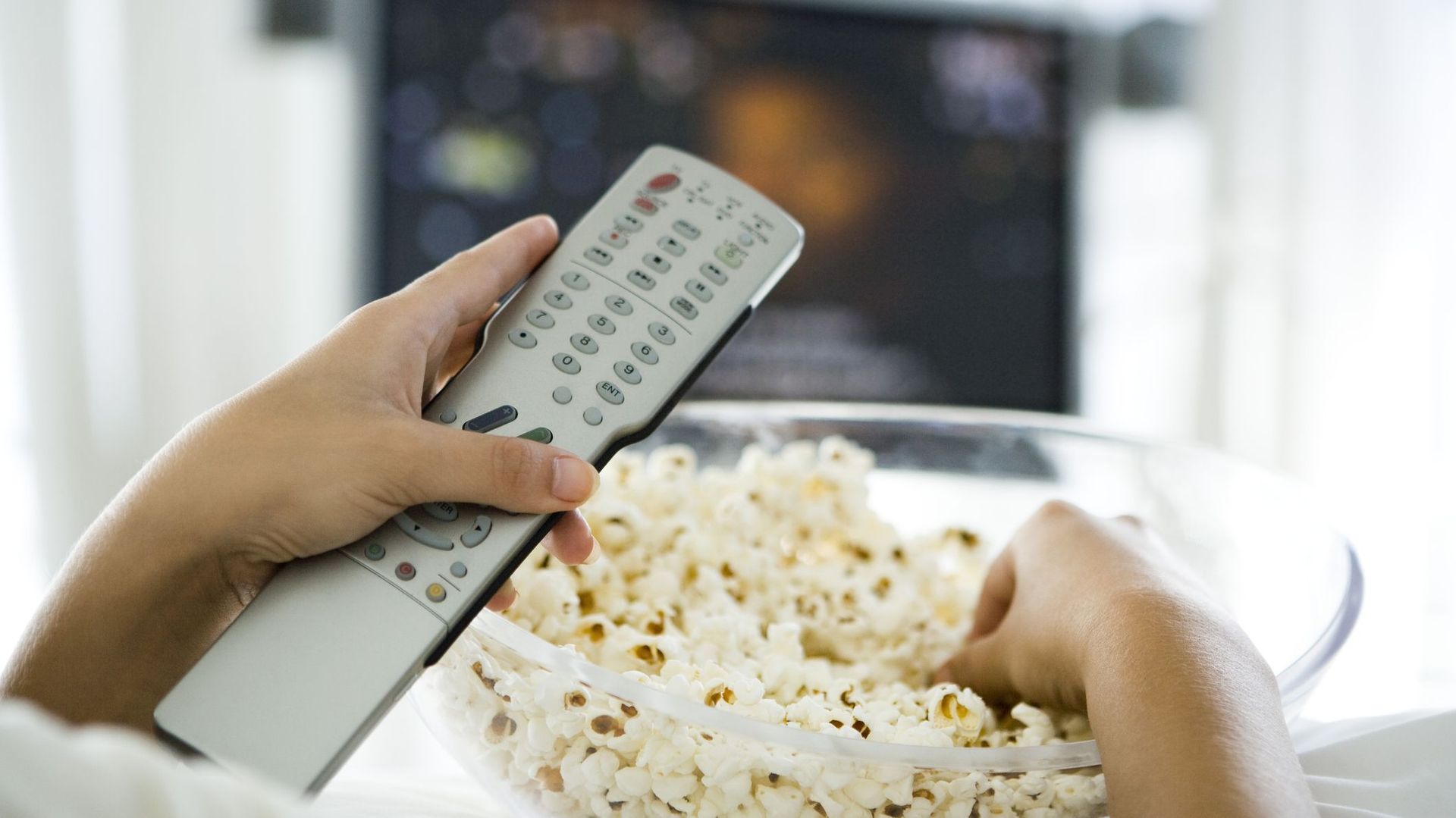 Person watching television, holding remote control and bowl of popcorn, cropped view