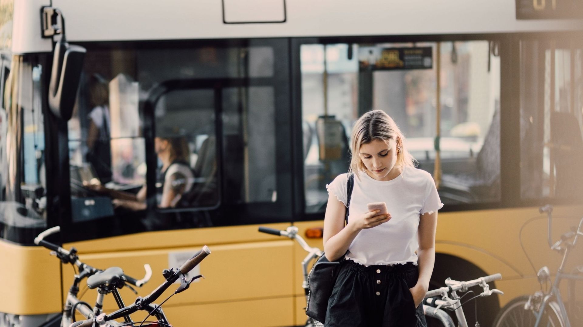 Young woman using smart phone while standing by bicycles against bus in city