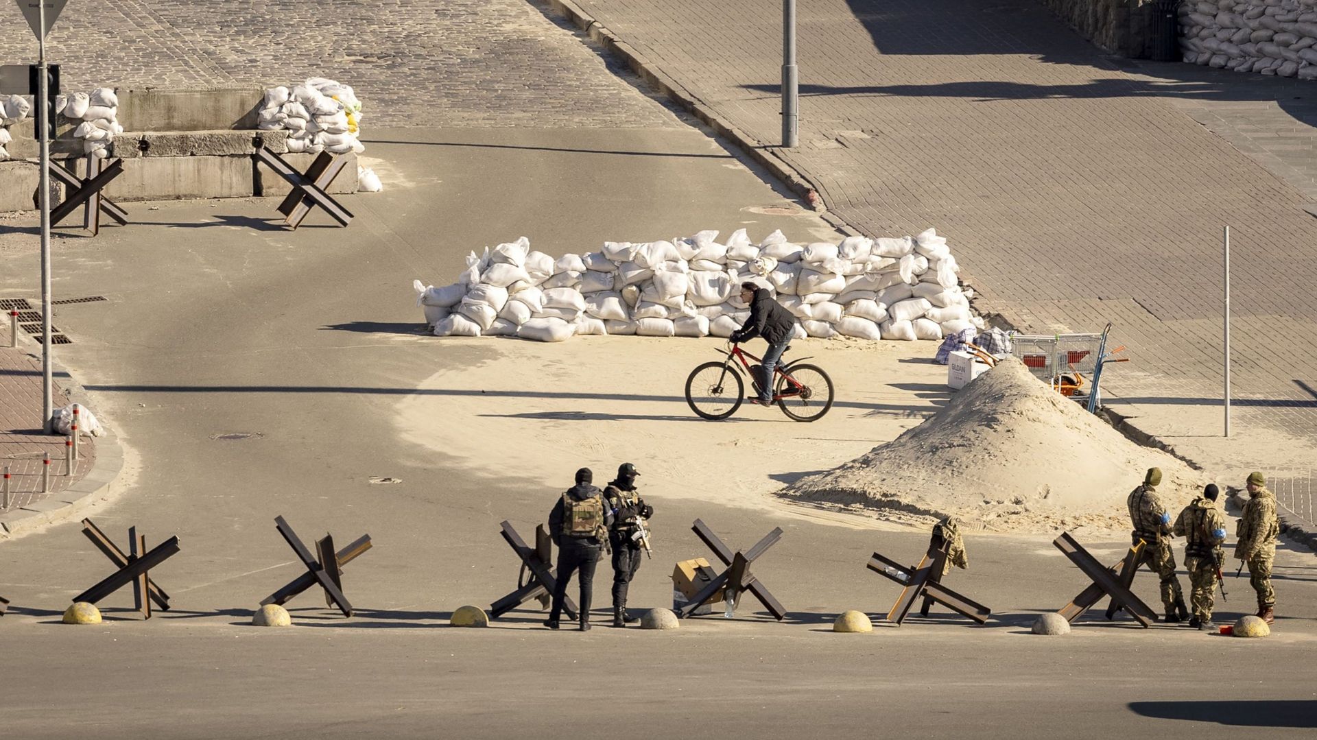 Un cycliste passe devant des militaires ukrainiens qui montent la garde à un poste de contrôle militaire dans le centre de Kiev, le 15 mars 2022, au 20e jour de l’invasion russe de l’Ukraine. Les délégations russes et ukrainiennes ont repris les pourparle