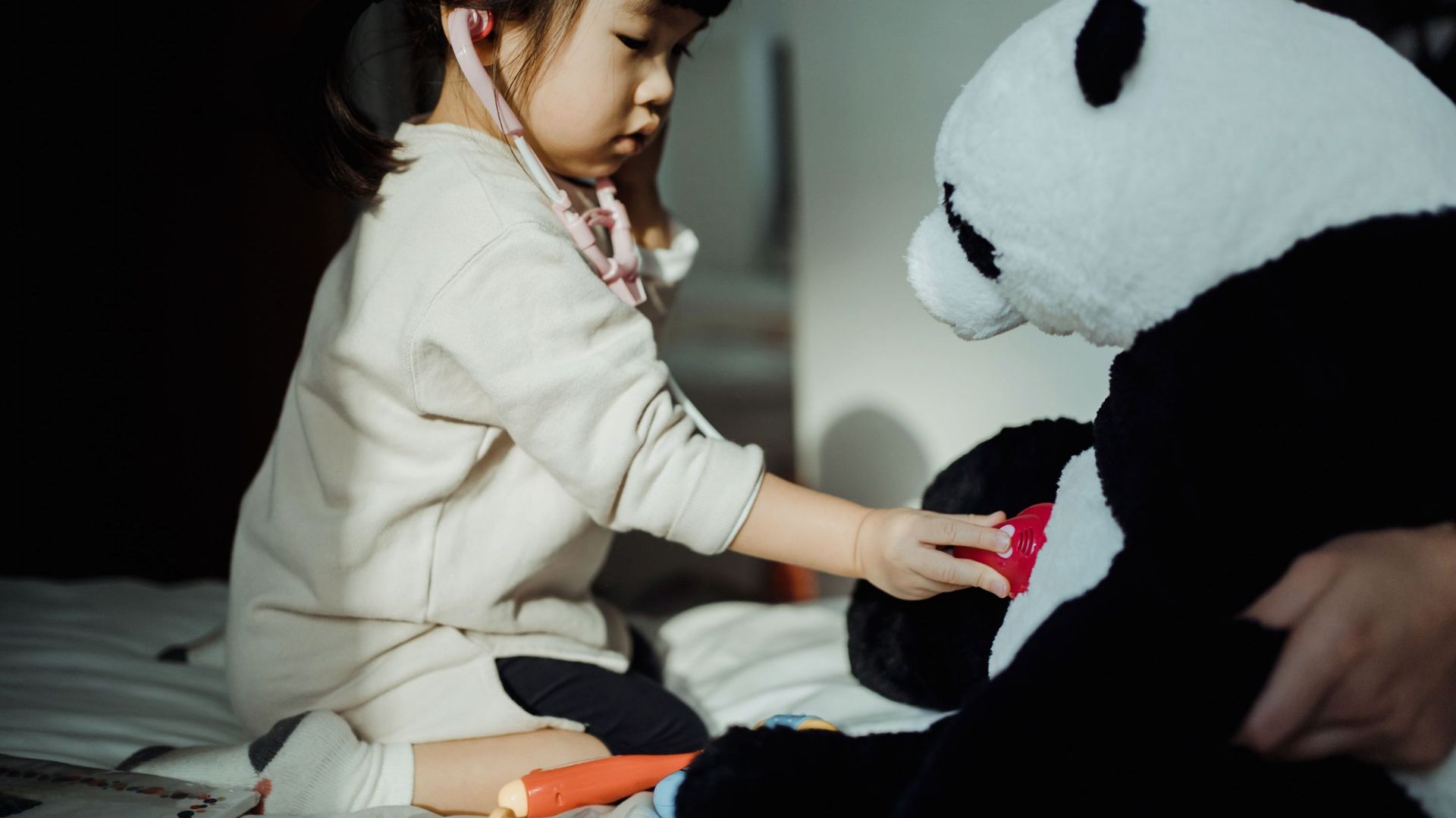 Adorable little Asian girl playing a doctor game with her beloved soft panda toy, listening the heartbeat of the soft panda toy with a stethoscope at home