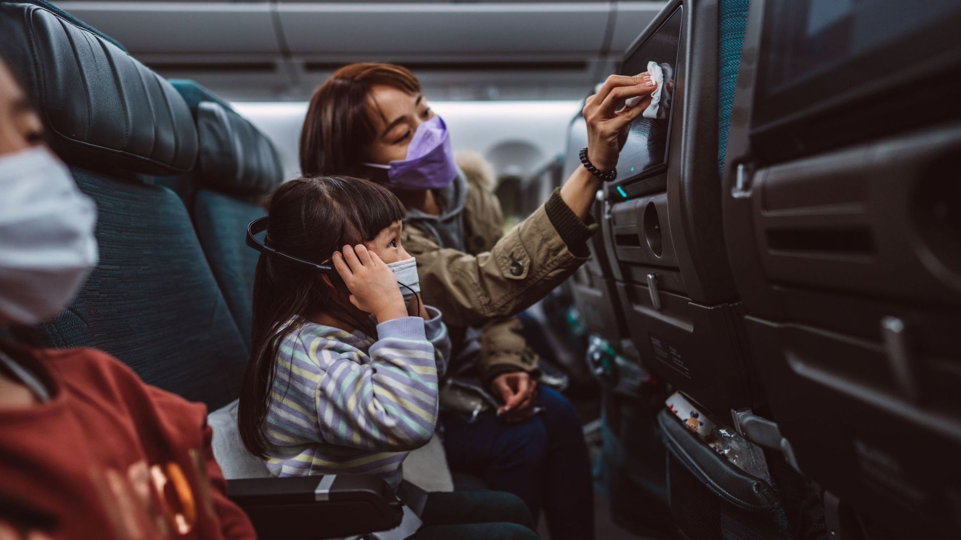 Mom &#38 ; daughter in protective face masks cleaning the seat-back TV screen with disinfectant surface wipe while travelling on the airplane
