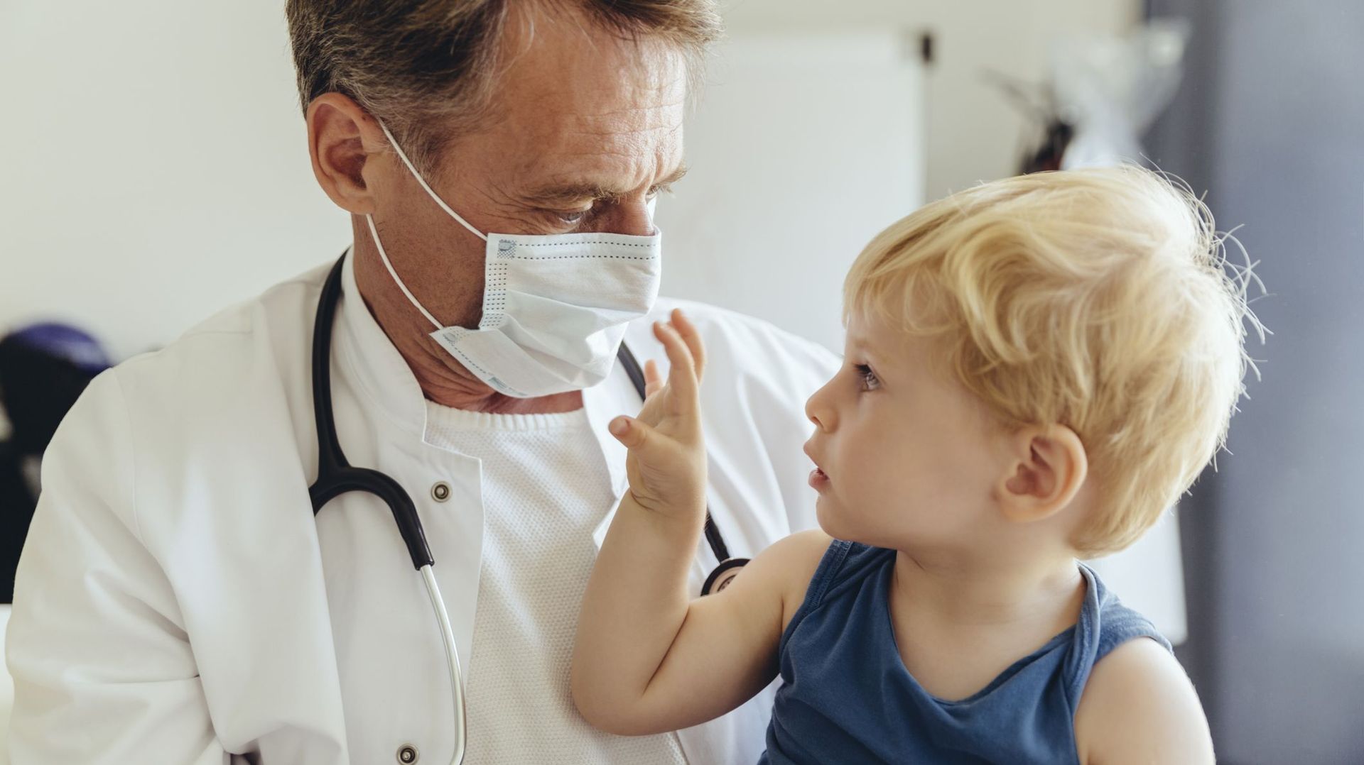 Toddler sitting on lap of pediatrician, wearing protective mask