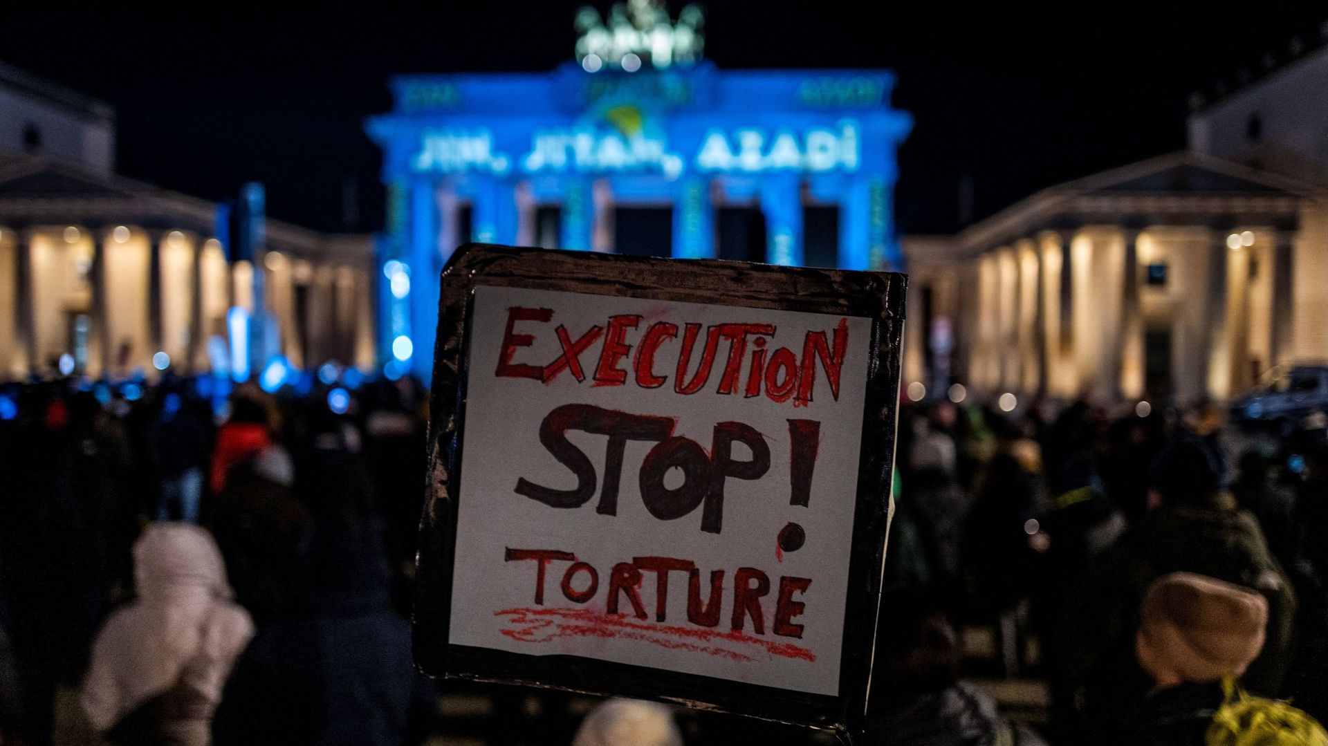 Un militant exhibe une pancarte portant l’inscription "Stop aux exécutions et à la torture", lors d’une manifestation de soutien aux manifestants en Iran, devant la Porte de Brandebourg illuminée des mots "Femme, Vie, Liberté" en plusieurs langues dont le