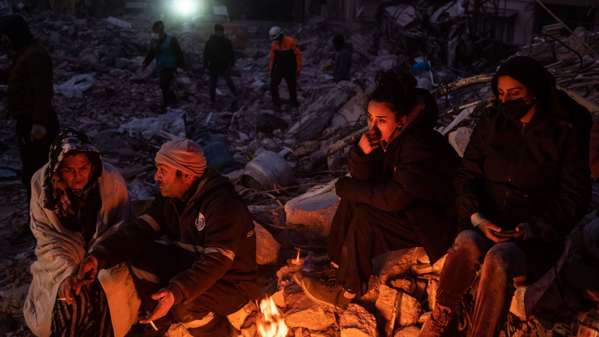 15 FÉVRIER : Des amis et des parents attendent près du feu en regardant une pelleteuse creuser un bâtiment détruit le 15 février 2023 à Hatay, en Turquie