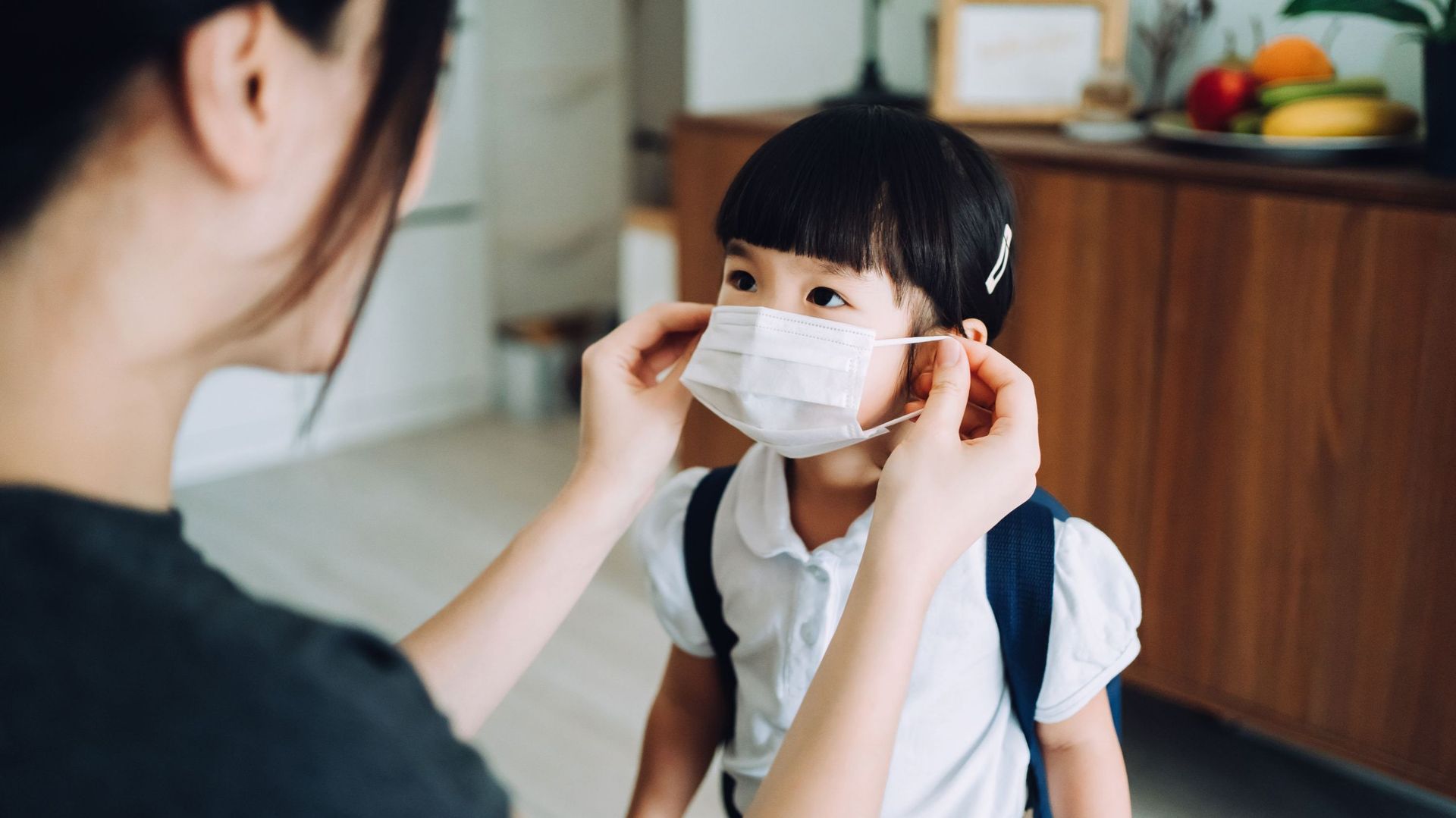 Young Asian mother preparing for her little daughter&#39 ; s first day of school and putting on a protective face mask on her daughter&#39 ; s face to protect and prevent from the spread of viruses