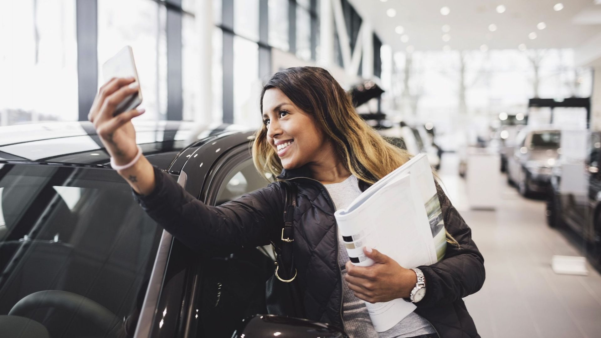 Smiling woman taking selfie with new car in showroom