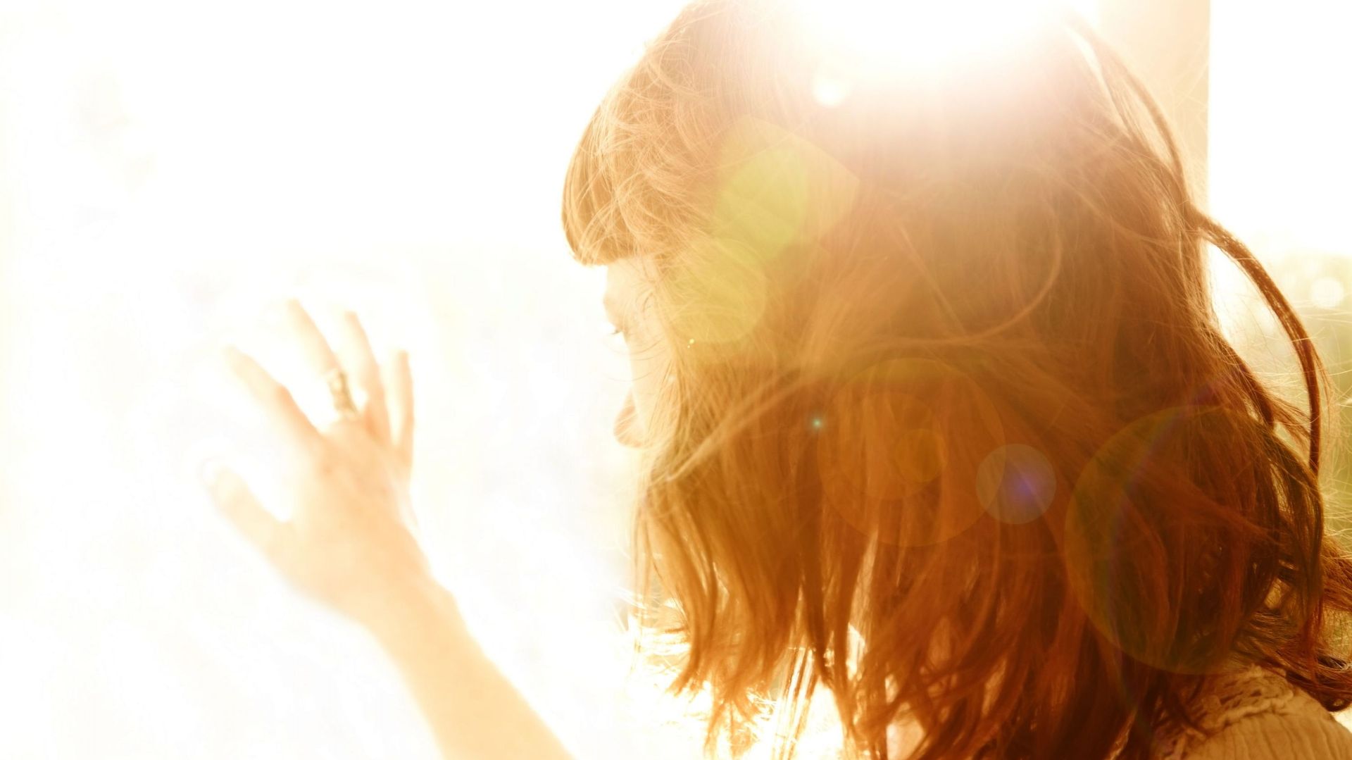Redhead woman backlit by sun with hand on glass window