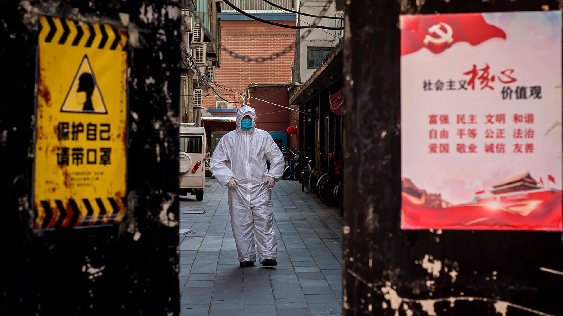 Un homme en tenue de protection à l’extérieur d’un bâtiment résidentiel où une personne a été détectée malade à Pékin. Un avis sur la porte d’entrée indique "protégez-vous", à côté d’une affiche de propagande du gouvernement.