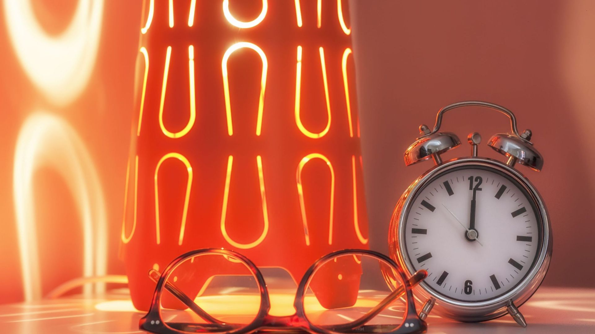 Alarm clock and glasses on a bedside illuminated by a modern lampshade