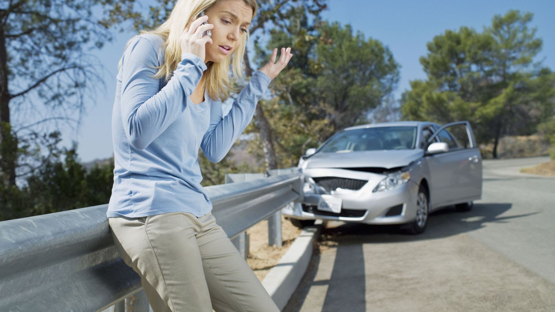 Frustrated woman using cell phone next to car wrecked on guardrail