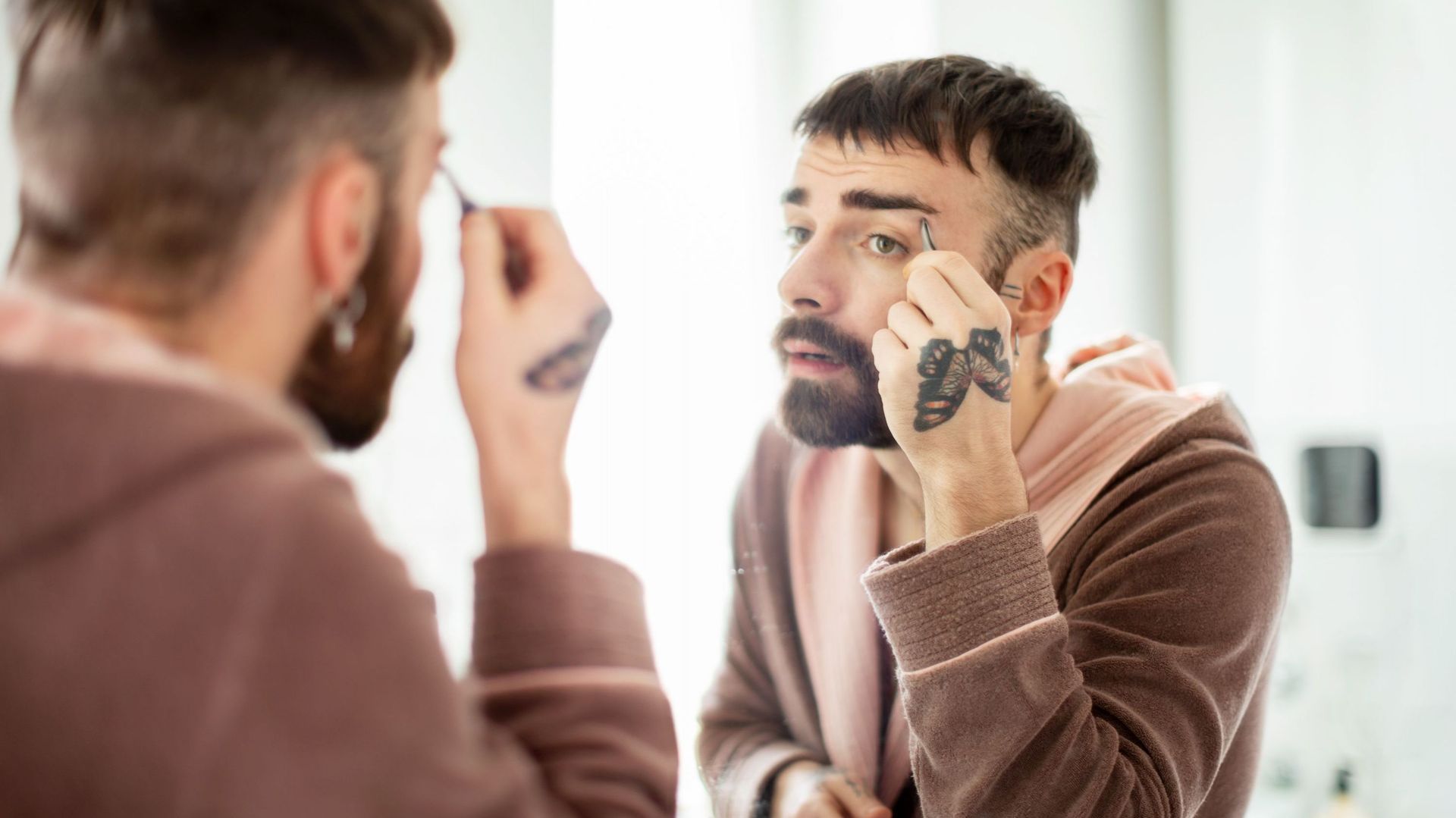 Young man wearing bathrobe plucking eyebrows in front of the bathroom mirror, butterfly tattoo on hand