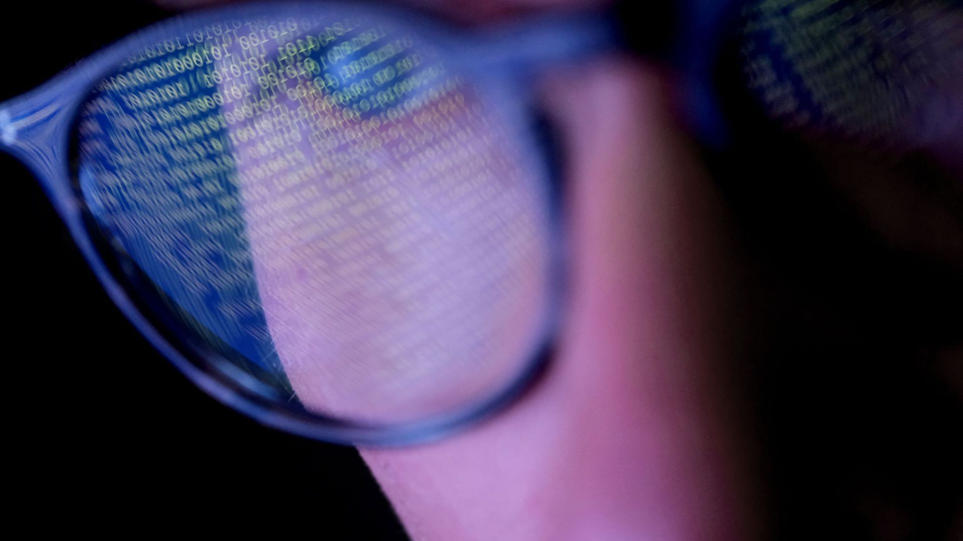 A person with glasses in a dark room looking at computer screen that&#39;s displaying codes or programming, with reflections on the glasses keeping the subject engrossed in