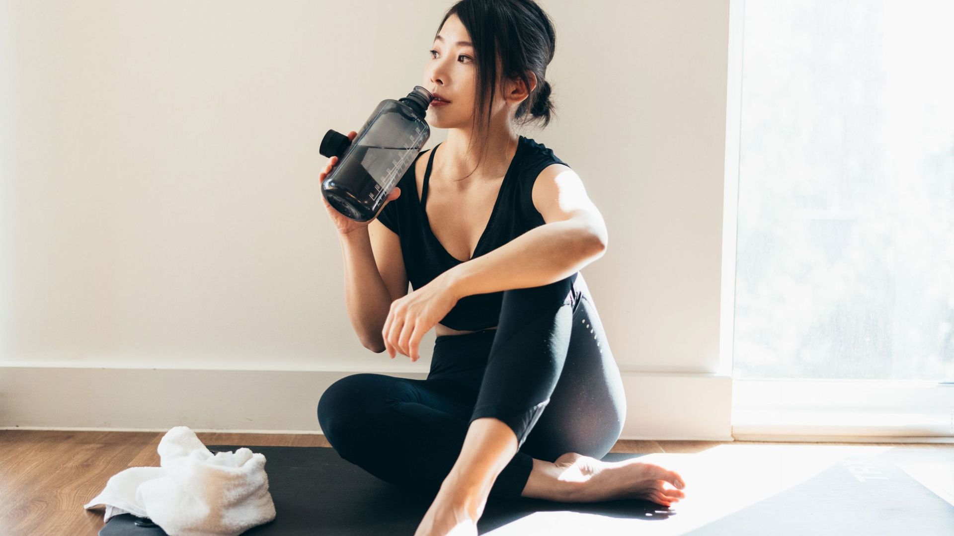 Asian woman drinking water with a water bottle, sitting on exercise mat after doing yoga