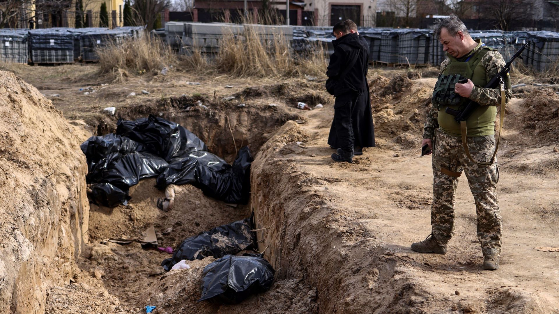 Un soldat ukrainien regarde des sacs mortuaires tandis que des prêtres prient devant une fosse commune dans les environs de l’église Saint-André à Bucha, le 7 avril 2022, dans le cadre de l’invasion militaire lancée par la Russie en Ukraine.