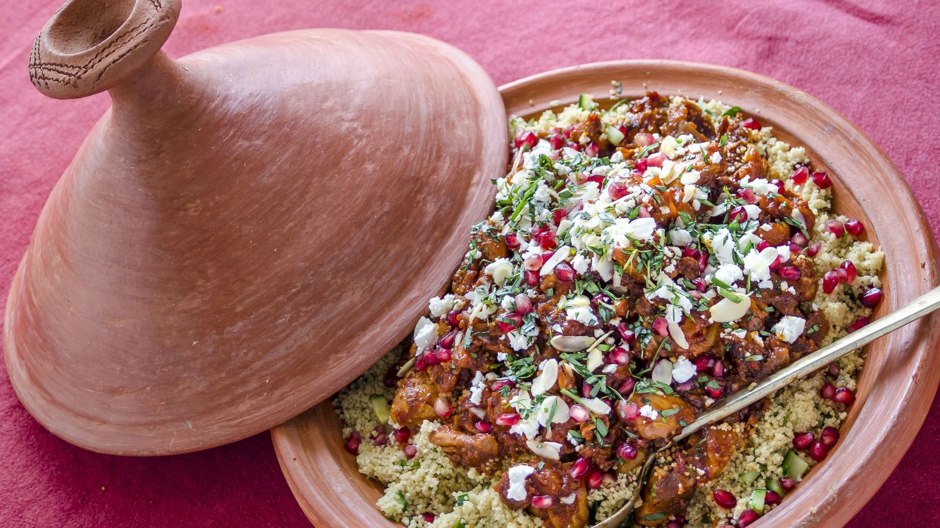 Chicken cous cous tagine with pomegranate seeds, feta cheese and herbs in a terracotta tagine with a serving spoon on a red tablecloth